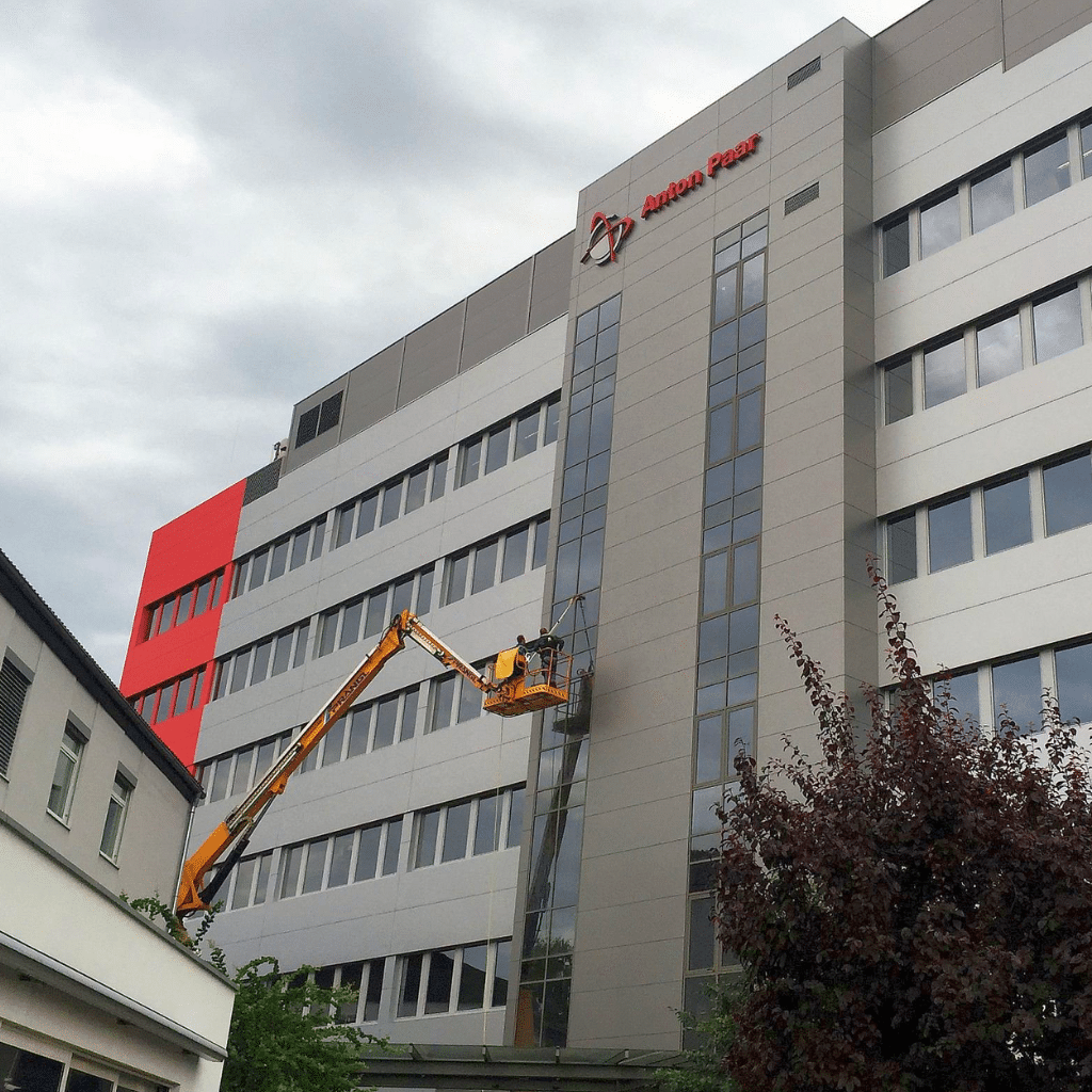 Ein Arbeiter reinigt die Glasfassade eines modernen, mehrstöckigen Bürogebäudes mit einer gelben Gelenk-Teleskopbühne. Die Fassade ist grau und weiß mit roten Elementen. Der Himmel ist bewölkt.