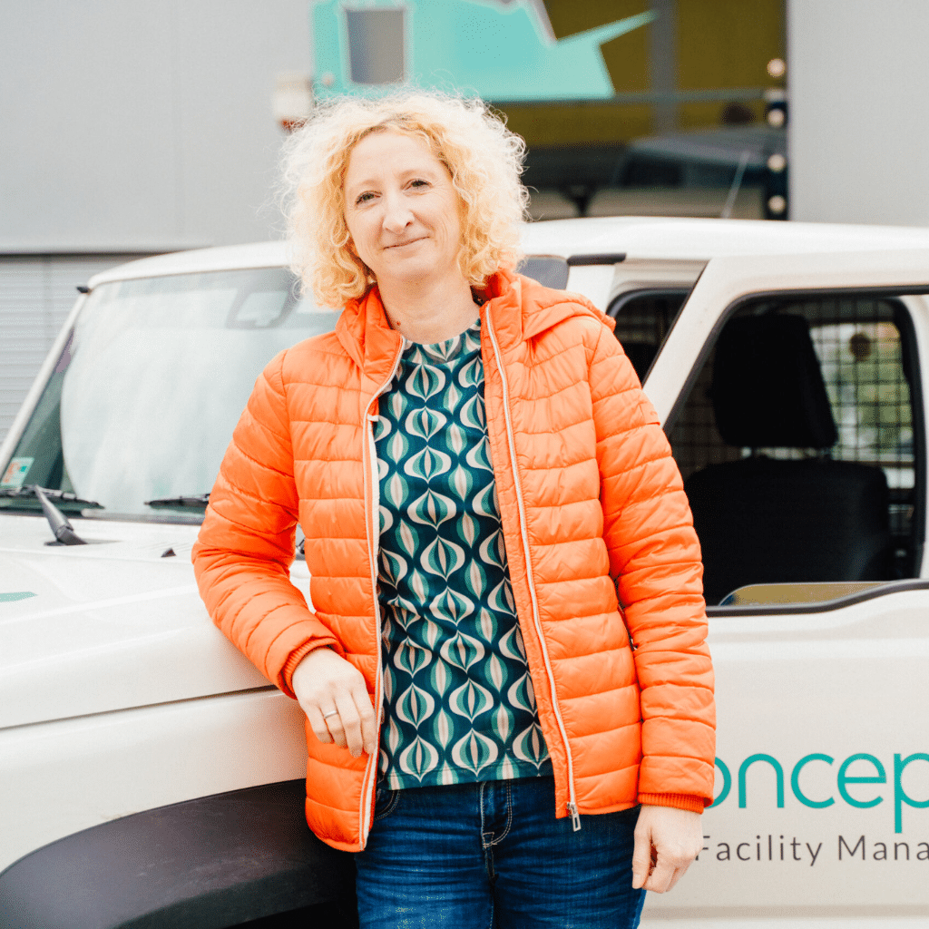 Eine Frau mit lockigem blondem Haar und oranger Jacke lehnt an einem weißen Firmenwagen mit der Aufschrift „Concepts Facility Management“.
