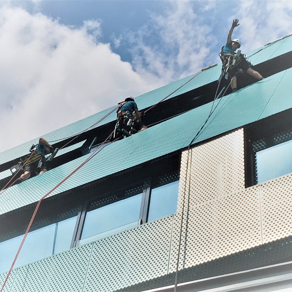 Drei Industriekletterer führen Fassadenarbeiten an einem modernen Gebäude mit grün und weiß perforierten Fassadenelementen durch. Sie sind mit Seilen gesichert und tragen Helme. Der Himmel ist teils bewölkt.